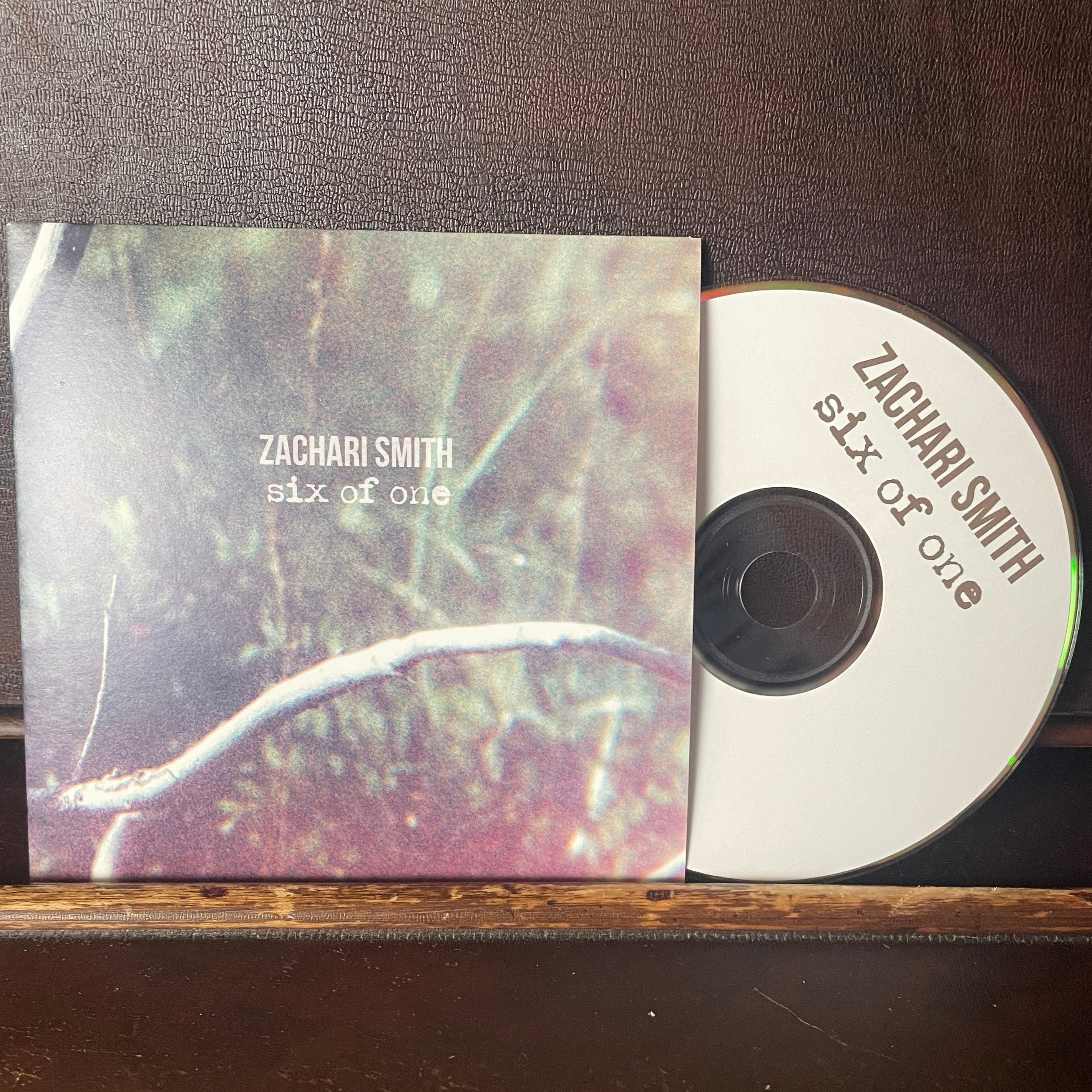 CD with a printed cover featuring 'Zachari Smith Six of One' on a wooden surface.